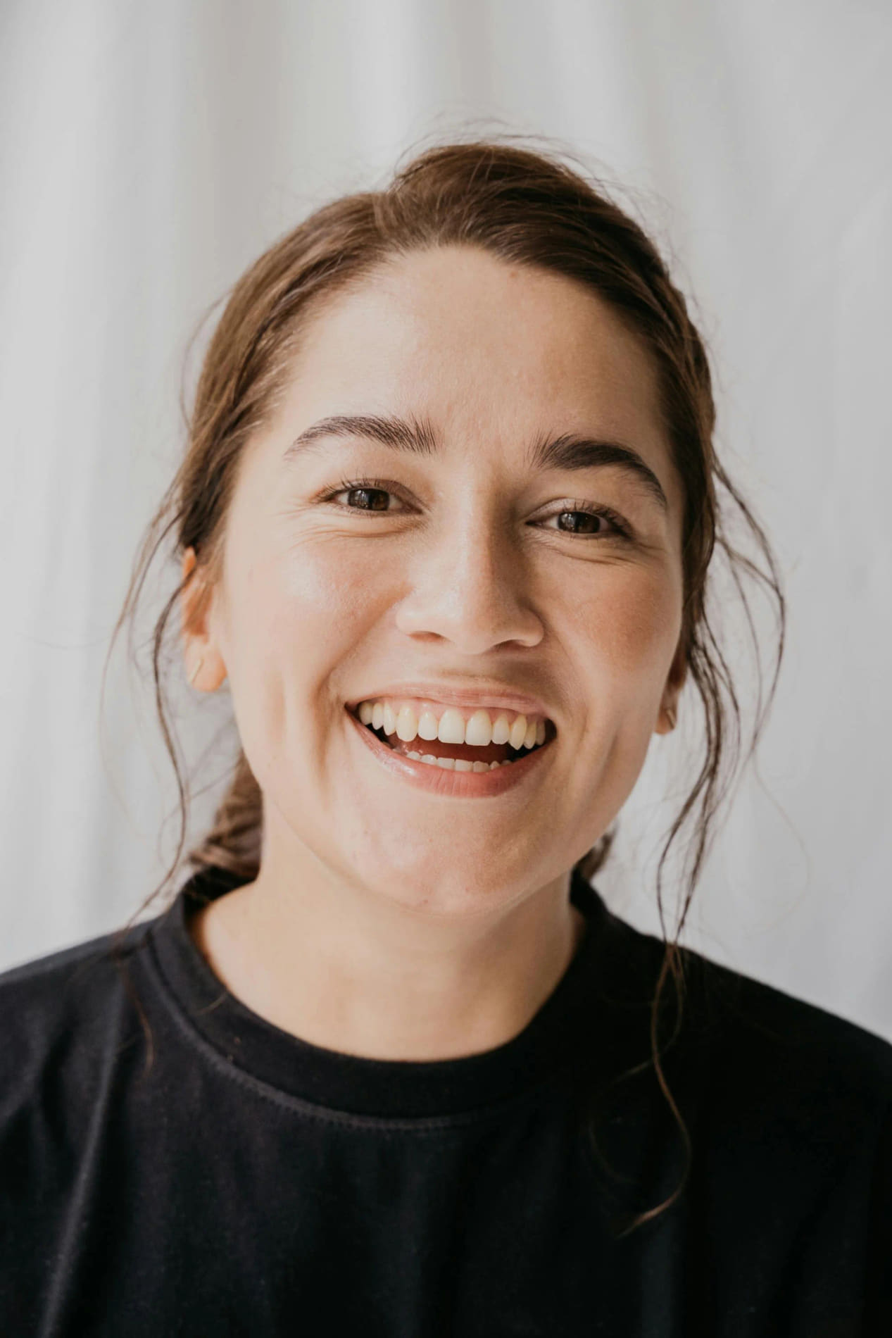 A close-up of a woman with brown hair smiling, wearing a black t-shirt against a plain background.