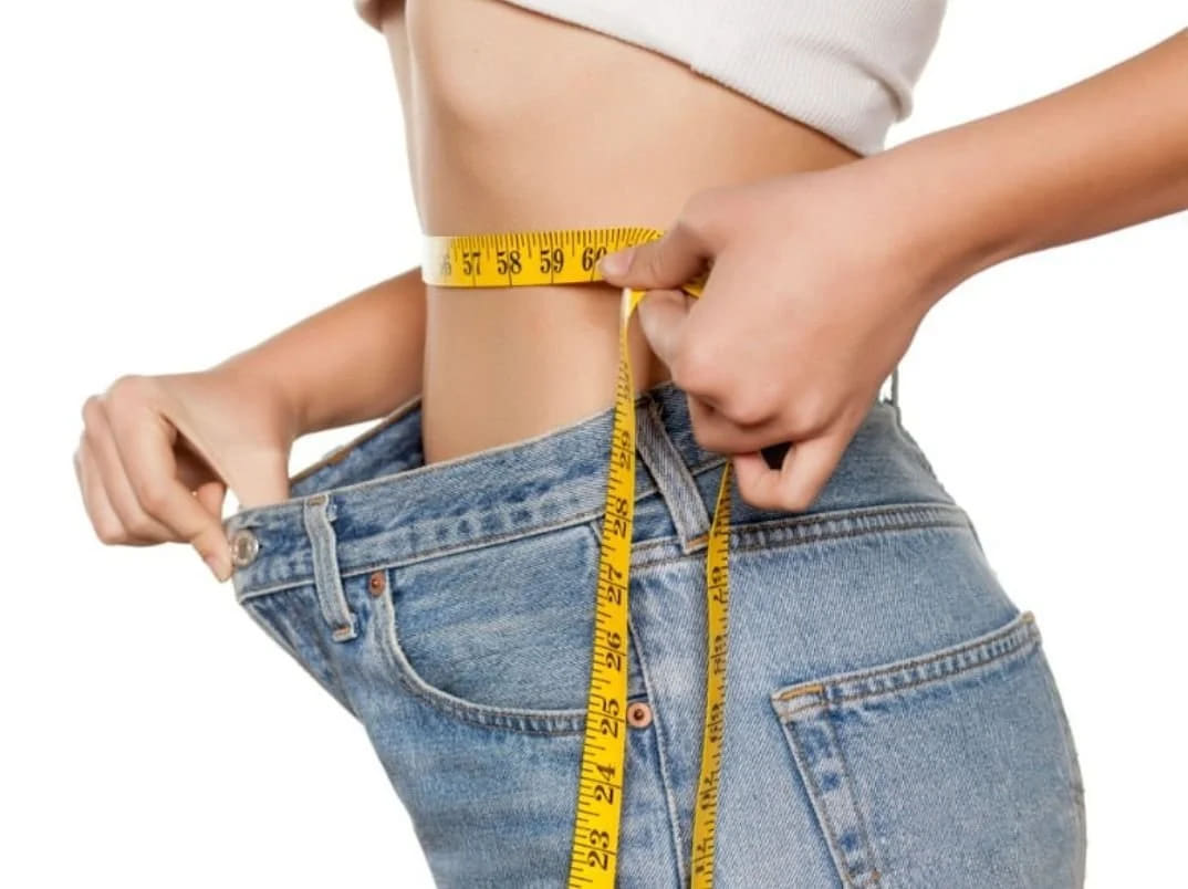 A person measuring their waist with a tape measure while wearing loose jeans and a cropped top.