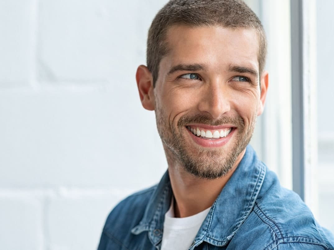 A smiling man with short light brown hair and a beard wearing a denim shirt, looking to his right near a window.