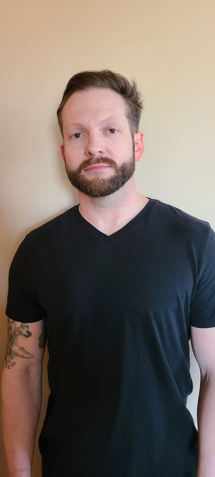 Portrait of Derek Thron with a beard and moustache, wearing a black V-neck T-shirt, standing against a beige wall.