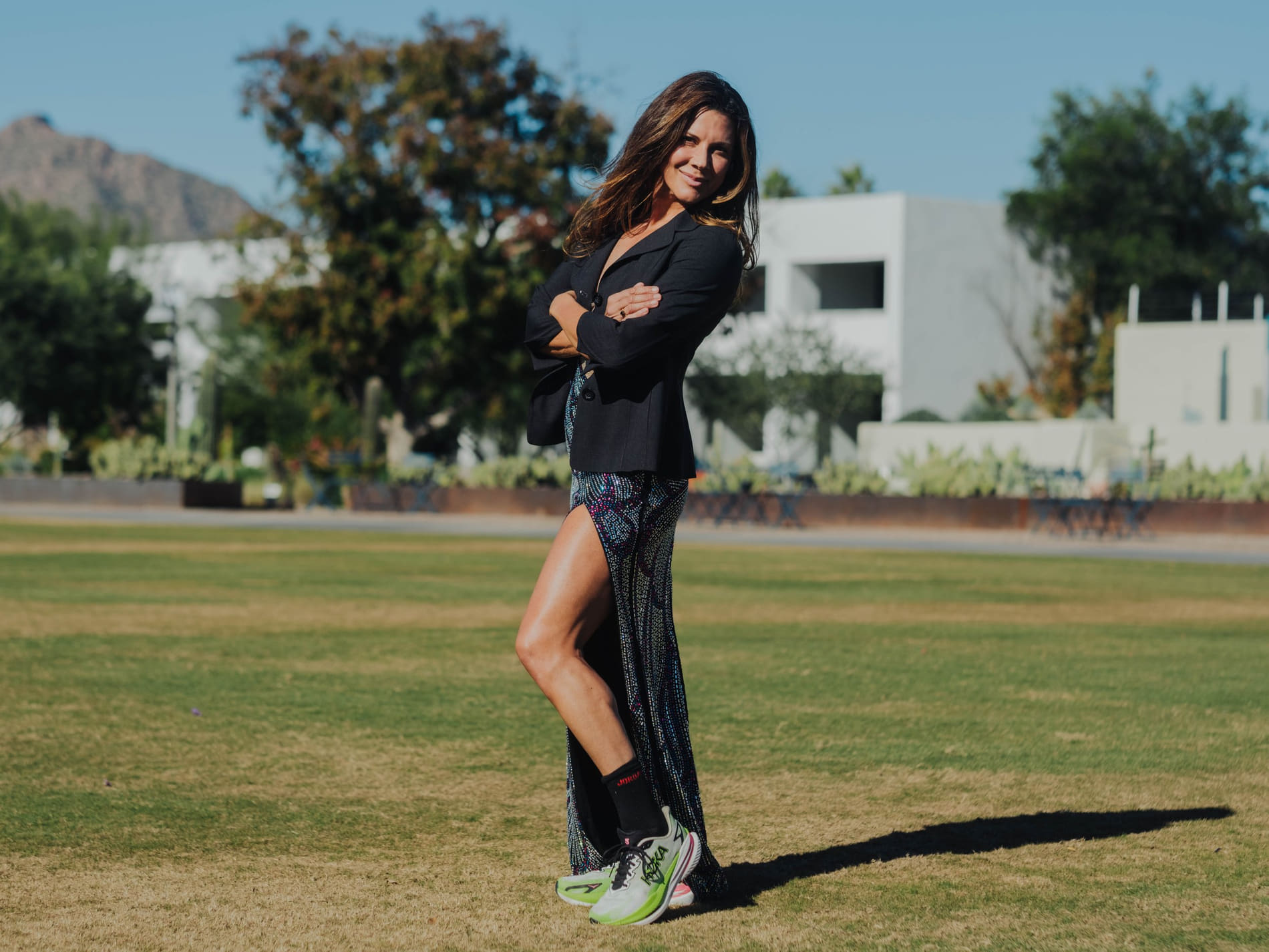 A woman standing outdoors on grass in a black blazer and patterned dress, arms crossed, smiling slightly.