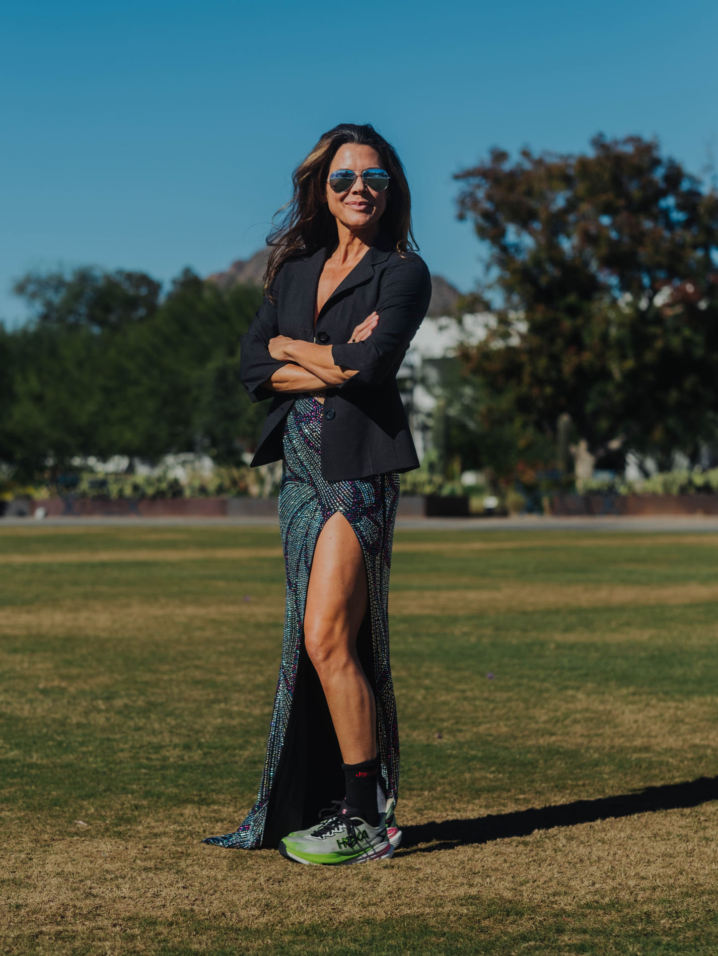Micole Neely standing on a grassy field in sunglasses, a black blazer and a long sequined dress, with trees and clear sky behind her.