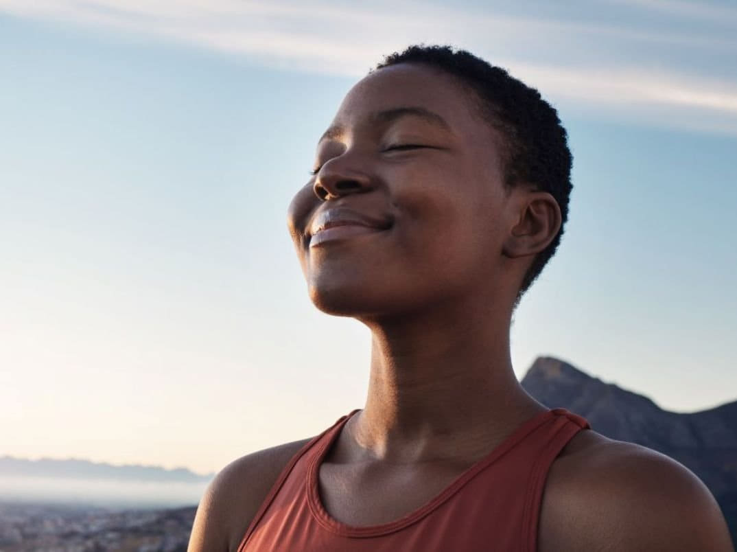 A woman with short curly hair, eyes closed, smiling peacefully outdoors with a mountain behind her.