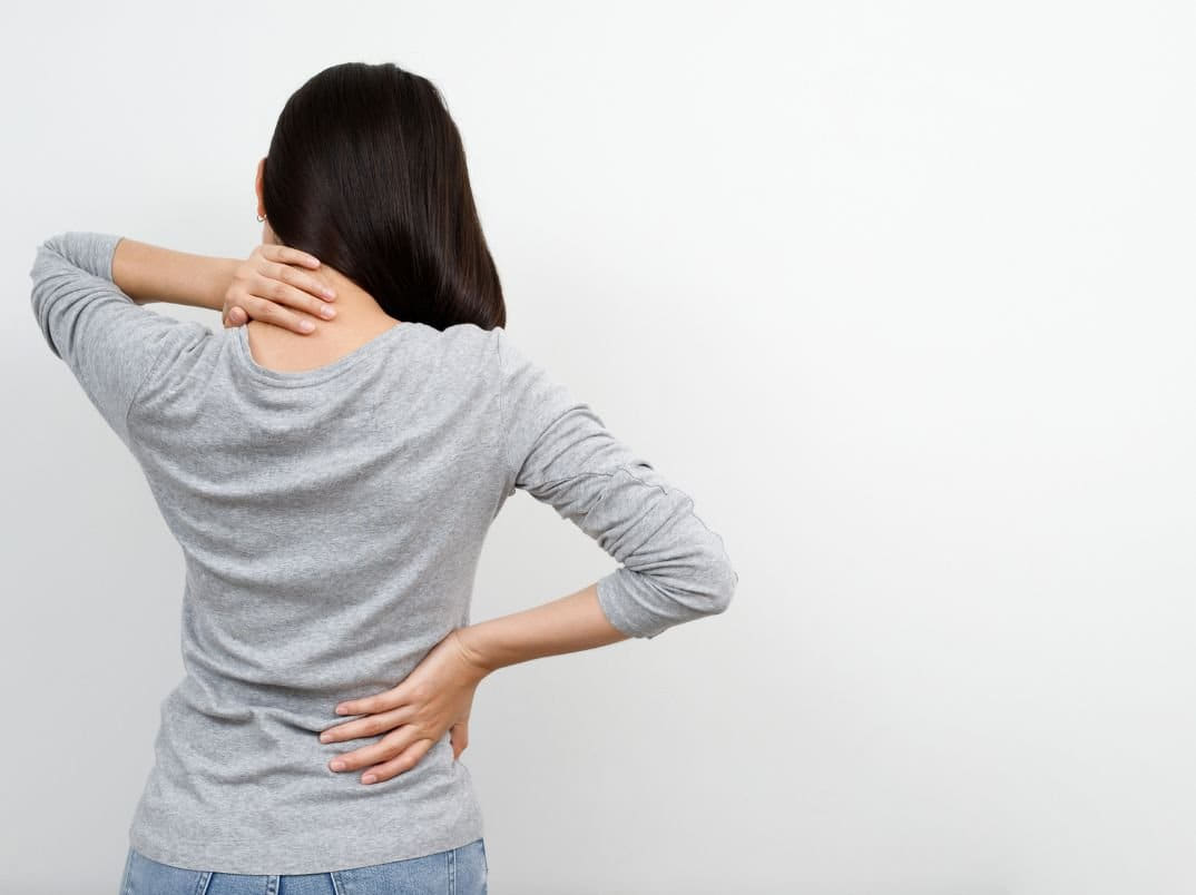 A woman in a grey long-sleeve shirt holding her neck with one hand and her lower back with the other.