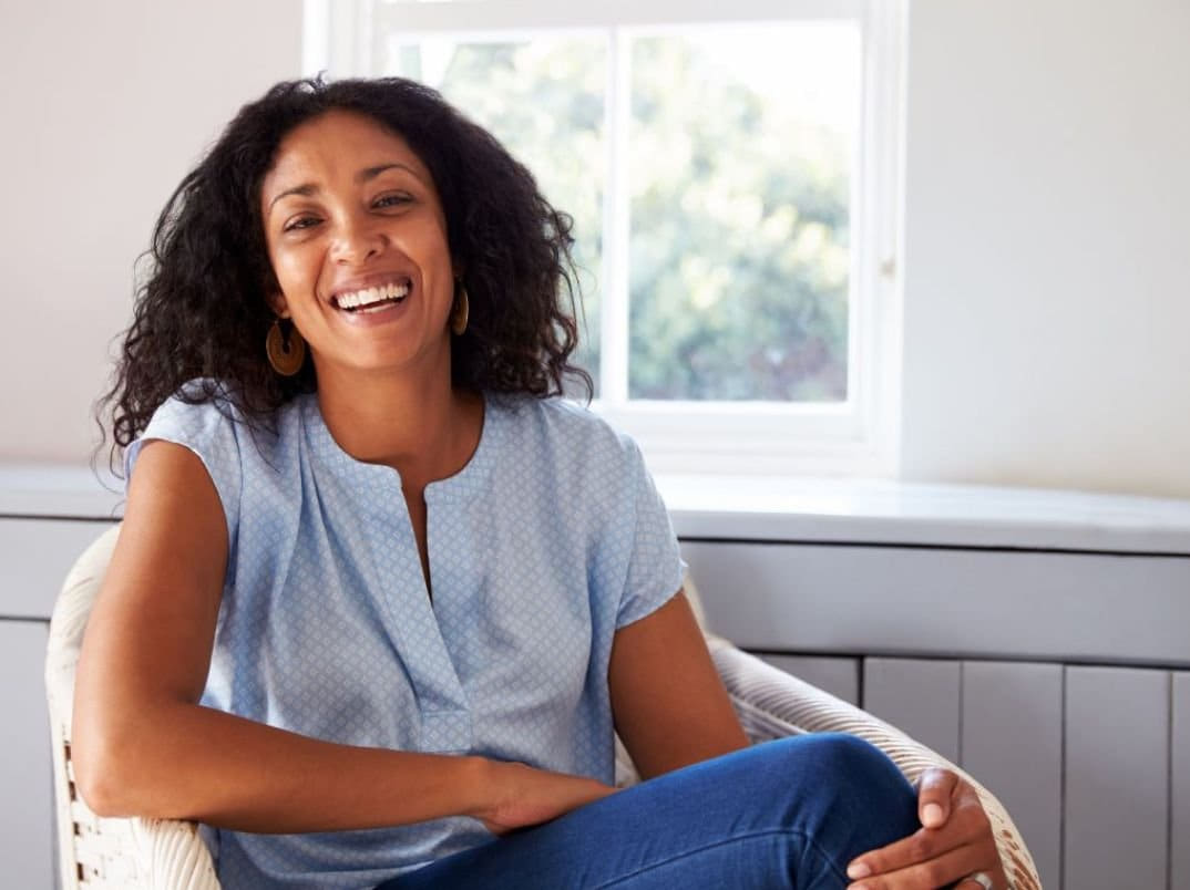 A woman with curly hair smiling and sitting on a chair in a well-lit room.