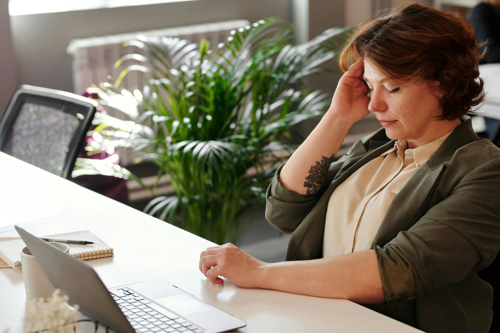 A woman with curly hair sitting at a white table holding her head with her hand, appearing tired, with a laptop and notepad in front of her.